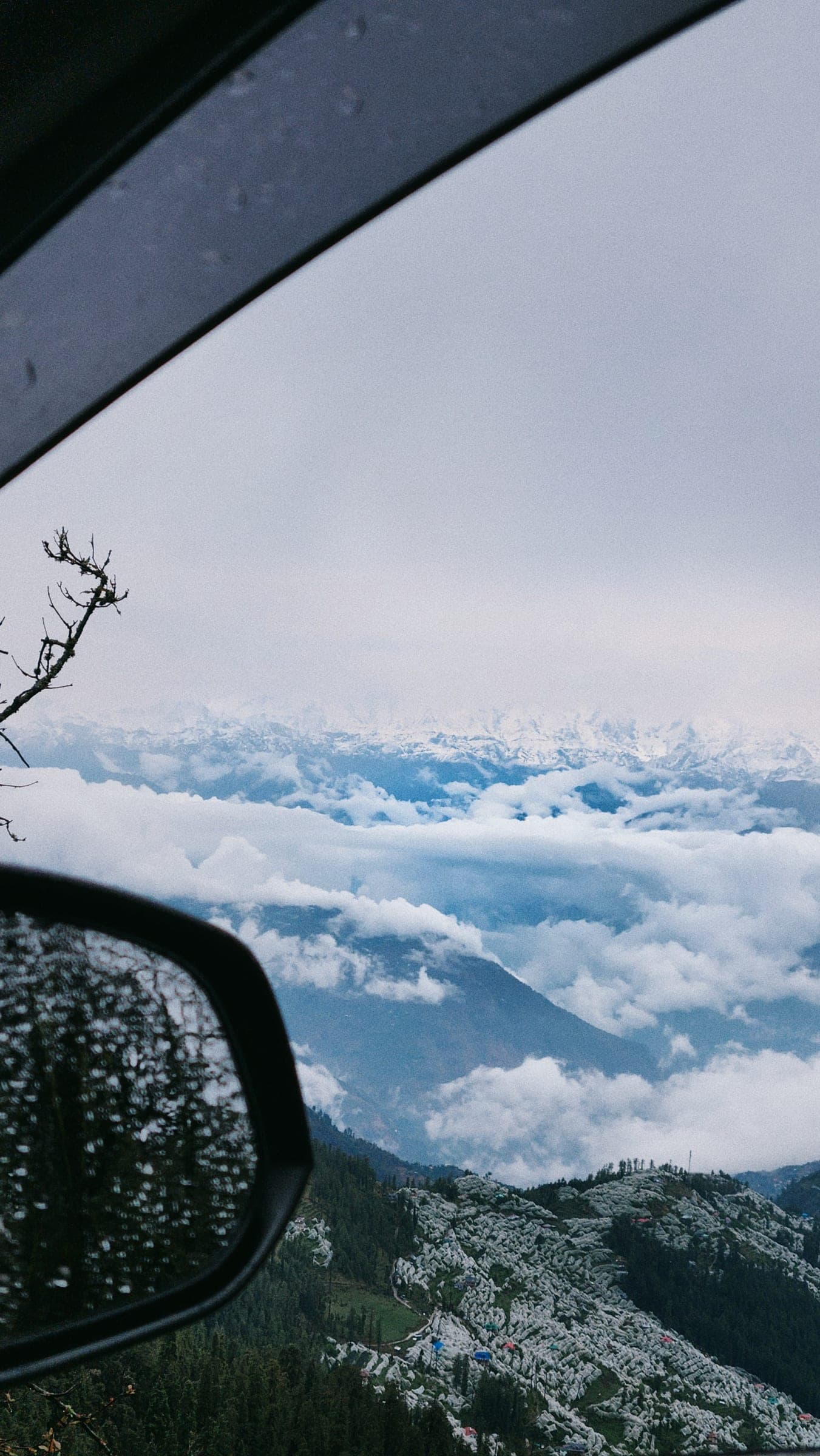 Shot from a car window: a wing mirror beaded with rain in the foreground, a sea of cloud filling the valley below and a far snow range on the horizon.