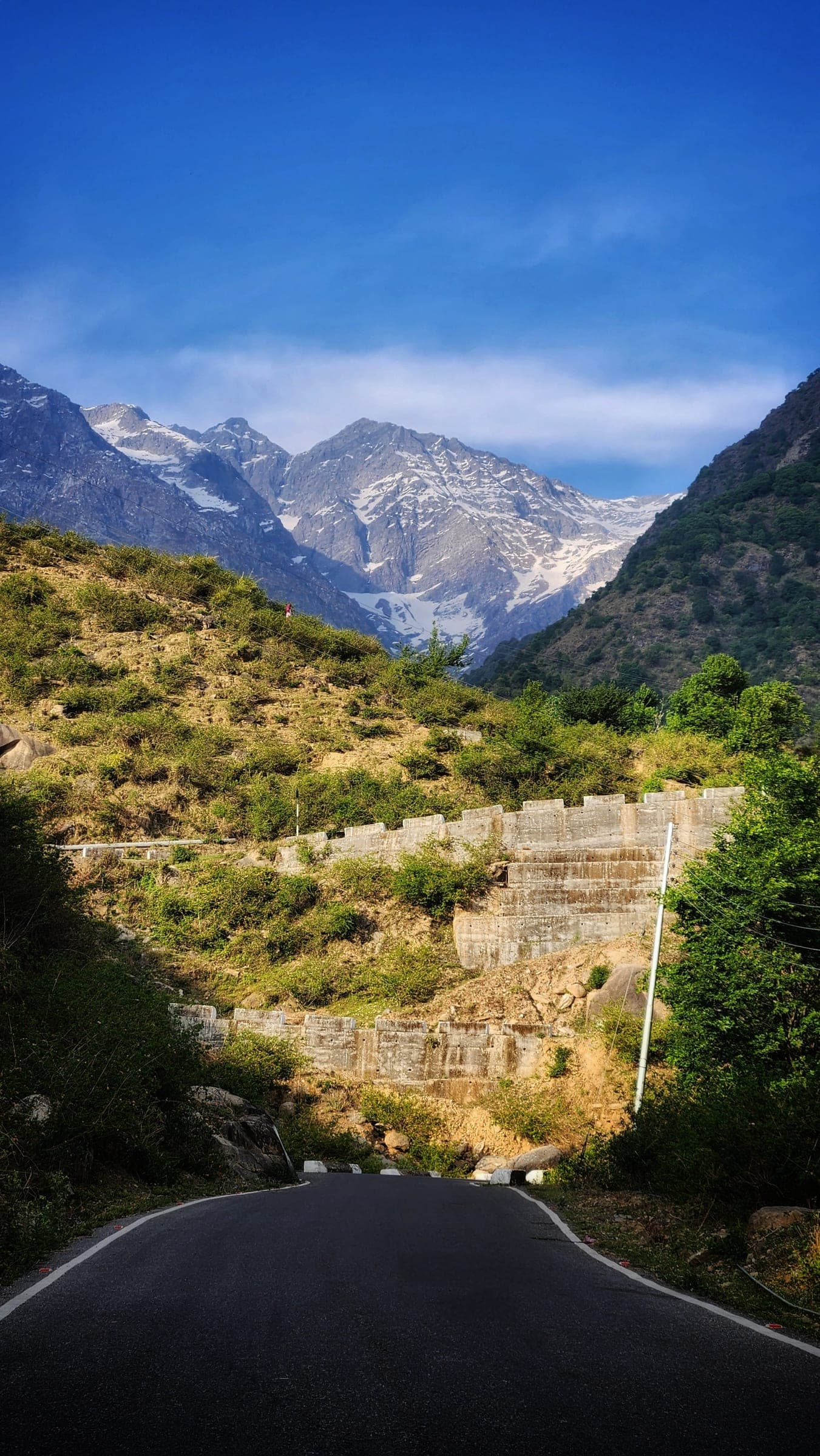 An empty road curving away toward snow peaks, green slopes and a stepped retaining wall on either side.