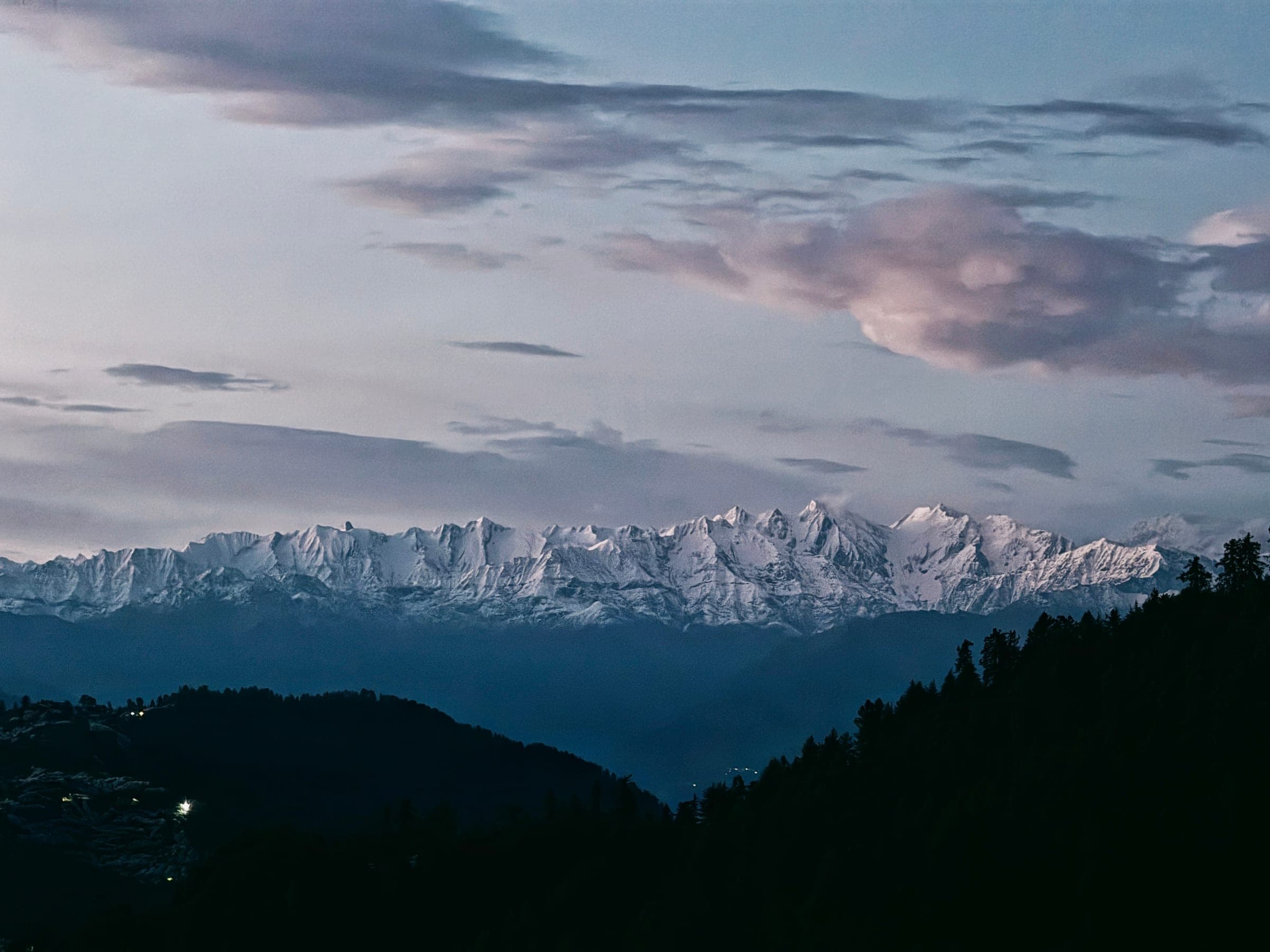 Blue hour: a long snow range along the whole horizon under banded cloud, forested ridges and a few village lights below.